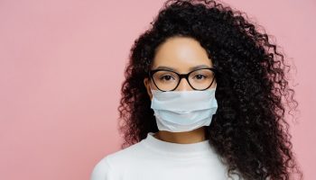 Covid-19, infectious virus. Close up shot of young woman with curly bushy hair, wears transparent glasses and medical disposable mask, cares about her health, protects in dangerious situation