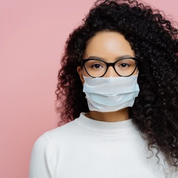 Covid-19, infectious virus. Close up shot of young woman with curly bushy hair, wears transparent glasses and medical disposable mask, cares about her health, protects in dangerious situation